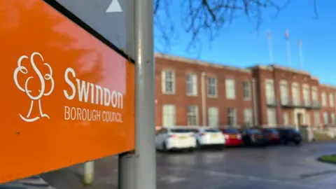 To the front left is an orange sign saying "Swindon Borough Council". In the blurred background are council offices.