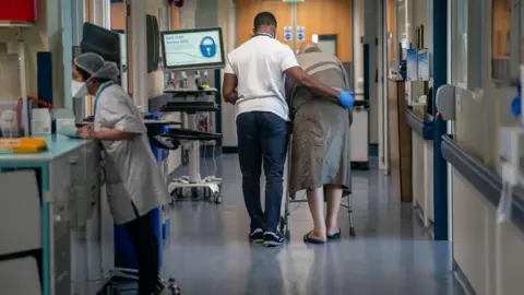 PA Media Nursing auxiliary helps an elderly patient with a zimmer frame to walk down a hospital ward