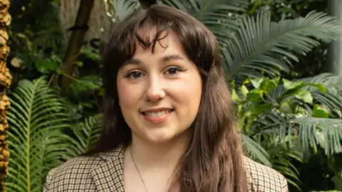 Margaret McDonald wears a brown checked blazer and stands in front of some large plants. She is smiling at the camera and has long brown hair and a fringe.