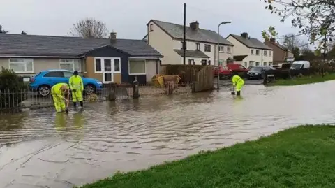 Three people in high-visibility clothing work to clear drains as a road is flooded in the village of Thursby. There is muddy water which is reaching homes. 