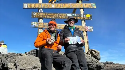 Two men dressed in mountain climbing gear posing at a mountain top with maps and signage behind them.