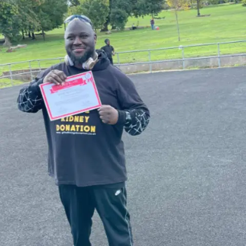 NHS A person standing on a park path holding a sign reading “Kidney Donation,” with trees and grass visible in the background.