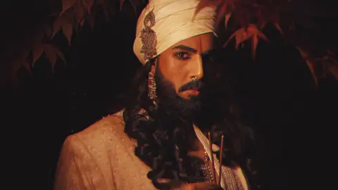 Rahual is wearing black kohl eyeliner and has a red sindoor mark on his forehead between his eyebrows. He has a white turban on his head with a matching white suit and is wearing silver and red jewellery on his ears and neck. He also has a silver septum piercing in his nose. Rahual has a black beard with a matching moustache and long black hair in ringlets. He is holding two lit incense sticks in his hand and looking at the camera. The background is black with branches of red autumn leaves hanging down in the top section of the photograph. 