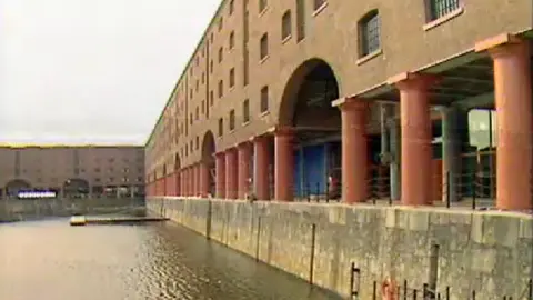 BBC Archives Albert Dock a restored brick building layout next to a full basin of water. The brick former warehouses have red pillars.