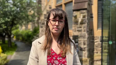 Dr Eileen Mitchell standing outside a grey and brone stone building, looking directly at the camera.  She has long, straight, brown hair with a fringe.  She is wearing glasses, a beige coat and a red patterned dress.  There is a tree-lined path behind her. 