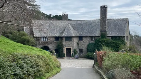 A large grey house sits at the end of the drive with the sea in the background.