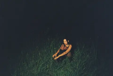 Natasha Ribeiro-Austrich Julia Wolf sits in a field at nighttime, surrounded by insects and tall grass, while illuminated by a mysterious light