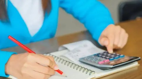 Getty Images Generic image of a woman in a turquoise cardigan and white t-shirt sitting at a table with a notepad, pencil, calculator and receipts working out her budget and debt.