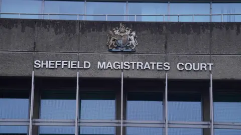 The exterior of Sheffield Magistrates' Court. The façade is constructed from grey concrete panels and has a stark, institutional appearance typical of civic architecture.
Prominently displayed across the centre of the building in large white capital letters is the text "SHEFFIELD MAGISTRATES COURT". Above the lettering is a decorative coat of arms mounted on the wall. The coat of arms features a shield surrounded by ornate heraldic elements, including animal figures on either side.