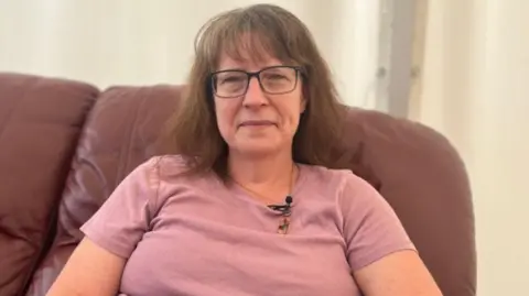 Karla Lee, wearing glasses and a pink t-shirt, sitting on a couch