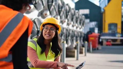 Angel Santana (via Getty) A woman with long straight black hair is wearing a hart hat and pointing to a laptop screen in a factory 
