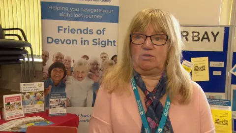 Mandy wears a coral coloured sweater, a purple scarf and glasses. She is stood in front of a Devon in Sight information board.