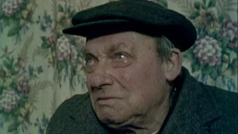 An elderly man in a pub, wearing a cap, with a tear in his eye.