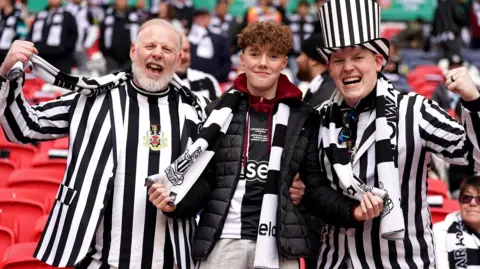 Three fans - two wear black and white suits. One fan has a black and white top hat. They are inside Wembley