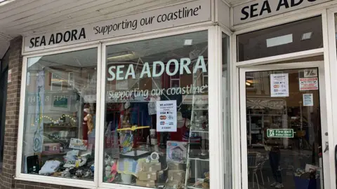 The front of Sea Adora which has a big window on the left and a door to the right. The words 'Sea Adora' is written on the window and below it has the words 'supporting our coastline'.