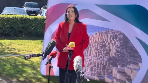 Shows a woman with long dark hair in red jacket and black dress standing in front of mics