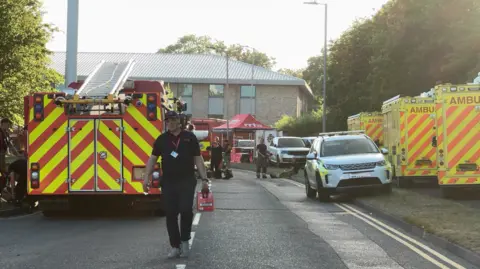 Fire appliances and ambulance vehicles line the approach road to an industrial building.