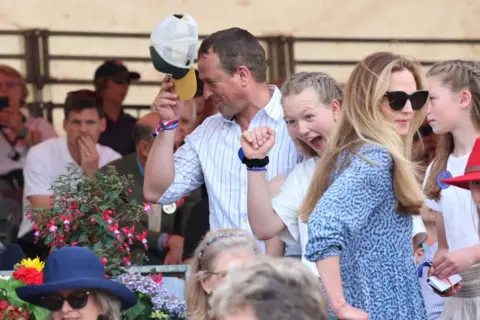 Chris Jackson/Getty Images Peter Phillips takes his cap off as Harriet Sperling stands in a blue dress and sunglasses a little lower down
