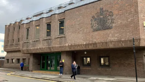 Chelmsford Crown Court, which is a red brick building on the side of a road. It has green doors at its entrance and windows spread out across the building. People are milling about outside.