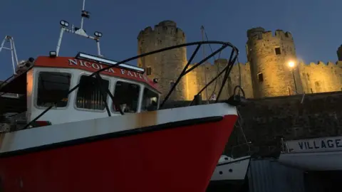 Family photo Part of a red fishing vessel is anchored at a harbour. It has the name Nicola Faith painted on it. Parts of a castle rampart are lit up in the background.