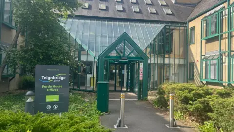 The front entrance of Forde House with a glass atrium. There are two metal bollards on the pathway to the entrance and a sign to the left.