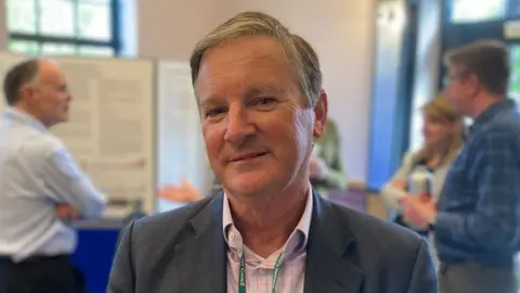 BBC Councillor Nick Botterill looks at the camera - it is a head and shoulder shot while at an event. He wears and grey suit jacket and pink check shirt, and has grey hair.