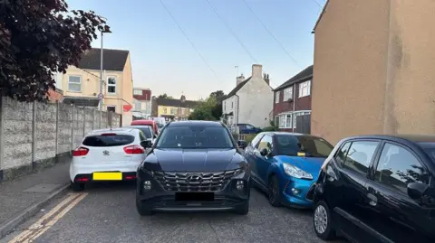 Cars can be seen parked on either side of a narrow street, with one black car trying to pass through in the middle.