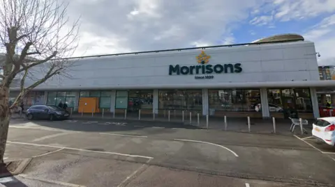 Google The Morrisons store pictured from the front. It is white and has Morrisons written in green on it. Cars are parked in the car park by the entrance and there is a tree with no leaves.