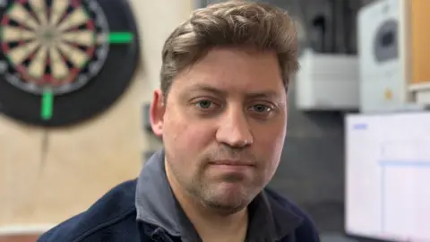 Ian Anderson has brown hair and is wearing a blue shirt. He is sat in front of a darts board and a blurred computer screen is also in the background.