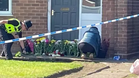 A woman kneeling down by the house and placing flowers by the door. A police officer stands to the left of the house.