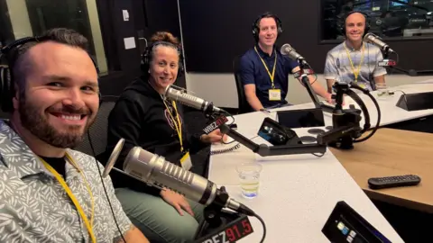 Elgin Police Department Three men and one woman, all wearing headphones, sit in a podcast recording studio with microphones and other equipment in front of them