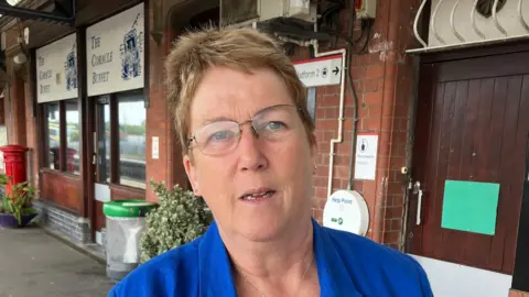 Ann Davies who has short light brown hair and glasses and is wearing a blue blazer