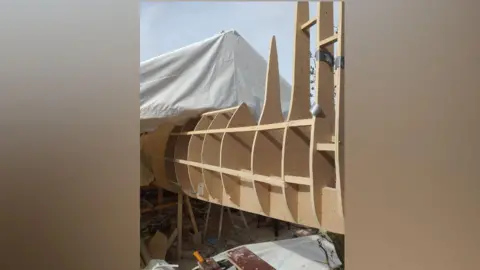 David Price The wooden tail of the Spitfire under construction poking out of a white marquee. It is surrounded by tools, sandpaper and building materials.