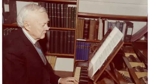 Armagh Planetarium A faded picture of an older man in a suit sitting playing a piano in front of a bookshelf.