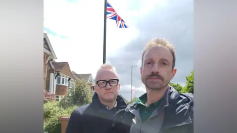 Two men stood in front of a lamppost with a Union Jack flag attached to it. Another lamppost in the background has a St George's Cross flag attached to it.