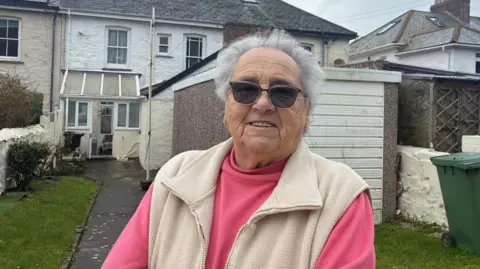 A woman wearing dark glasses smiles at the camera , she is standing in her back garden which is right next to the building site.