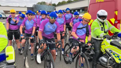 James Grant/BBC A peloton of bike riders dressed in pink bike jerseys with blue helmets lined up ready to cycle away. They are flanked by two police officers on motorcycles in high visibility clothing. 