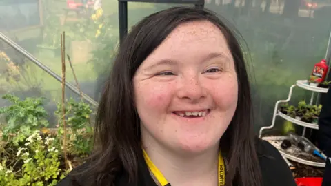 Savanna is smiling and looking into the camera. She has dark brown hair and is wearing a black top with a yellow lanyard over her neck. She's standing beside a greenhouse with plants behind her.
