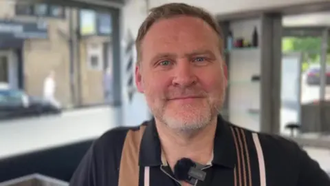 A picture of Jason Taylor, who runs a barbers in Brighouse, who has brown hair and a grey beard, you can see the windows and shelves of his barber shop behind him.