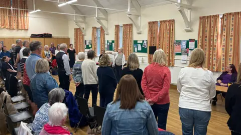John Devine/BBC People standing in rows and sitting on chairs, facing the front. They are in a community hall which has several windows with closed curtains.
