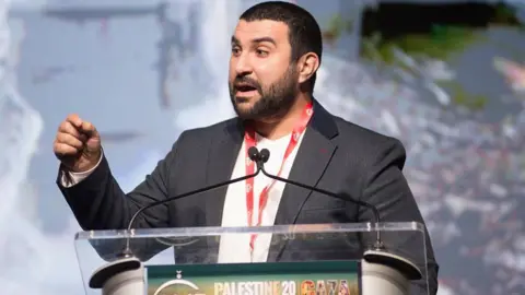 Hamdi speaking at a podium with his hand out to illustrate a point