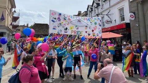 A group of people wearing pride flags and coloured polo shirts holding up a banner covered in ribbon and rainbow handprints, that says "Chippenham Pride"
