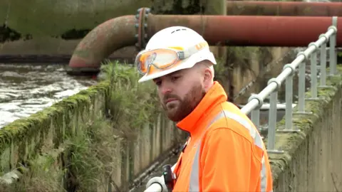 Ben Schofield/BBC An Anglian Water employee inspecting the sewage works at Bedford Water Recycling Centre. He is wearing a white hard hat, which has a pair of goggles laid over the top, and an orange high visibility jacket. He has a closely cropped beard and is looking down and to the left of the image. He is standing against a guard rail, beyond which is a body of water, concrete walls and in the distance behind him a large, industrial looking red pipe. The pipe runs from the top right of the image, behind the worker's head and then curves and disappears into the water. The concrete looks old and has moss and grass growing out of it at various points.