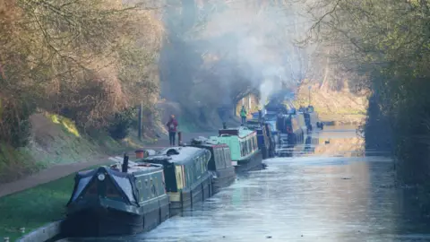 PA Media Several canal boats parked up in a line on the canal in a wintry scene with the water partially frozen over, the sunlight in the distance, and bare trees lining the bank