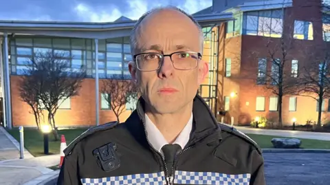 Paul Sanford is standing in front of the Norfolk Police and Fire Headquarters which is a large red bricked building with several glass windows. Sanford is wearing a white shirt, black tie and a police force jacket. He has short grey hair and is wearing clear and black framed glasses.