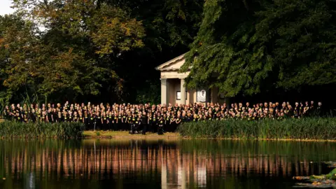 Lake in the foreground, which is reflecting the rows of singers wearing black T-shirts with yellow logos who are standing in front of a stone memorial with columns.  There are trees behind them.