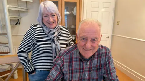 Stuart Porthouse sitting down smiling into the camera. He has white, short hair and is wearing a blue, white and red checked shirt. His wife is standing behind him with her hands in her pockets. She has white, chin-length hair with a fringe and is wearing a black and white striped long-sleeved top and a scarf. They are in a house with beige walls and wooden flooring. There is a white spiral staircase in the left corner.