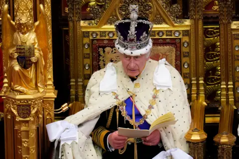 PA Media King Charles sits on a golden throne wearing the crown jewels, reading a speech