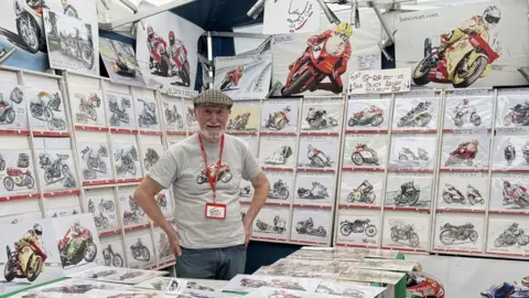 Hancox Art John Hancox, who is wearing a grey T-shirt with a drawing of a motorbike on it, surrounded by lots of examples of his work, which is also drawings of motorcycles.