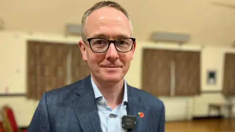 A man with short, fair hair with glasses, wearing a light blue shirt and dark blue suit